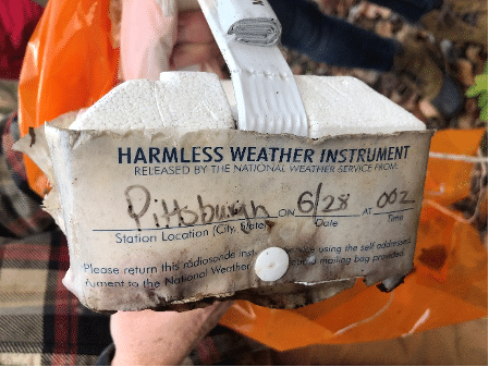 A photo of a deformed and probably dampened instrument package used by the National Weather Service to measure atmospheric variables aloft, after it fell to the ground. Writing on it's side states "Harmless Weather Instrument." 