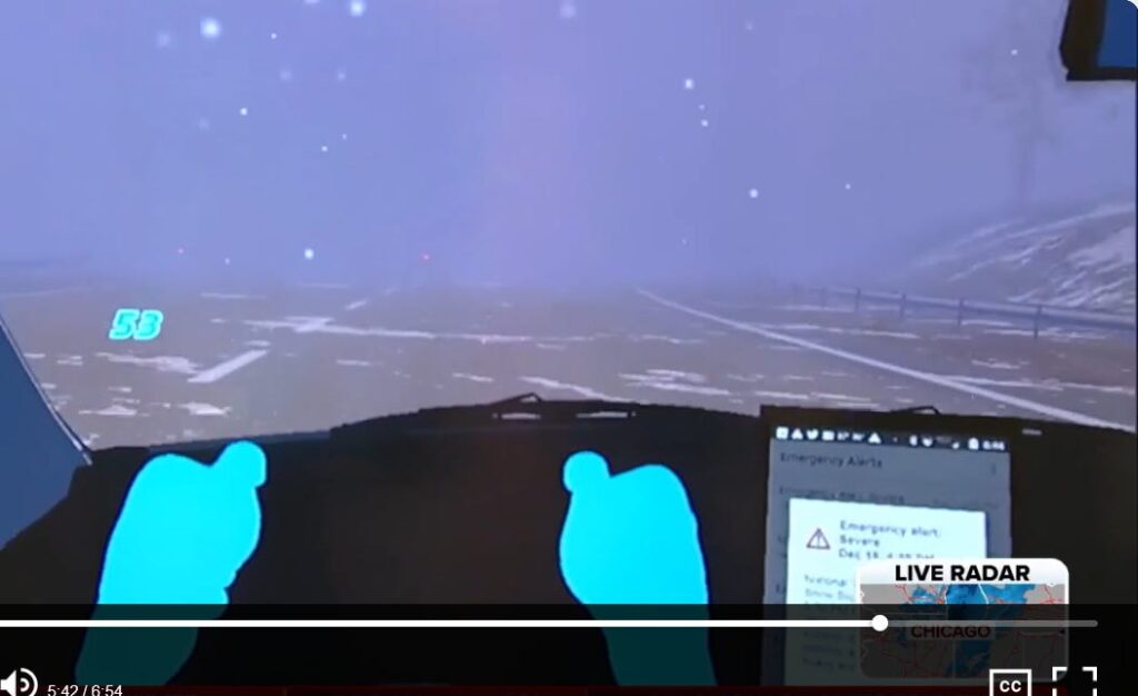 A firsthand image from the driving simulator, showing simulated hands on a steering wheel and the view of snowy conditions through the windshield.