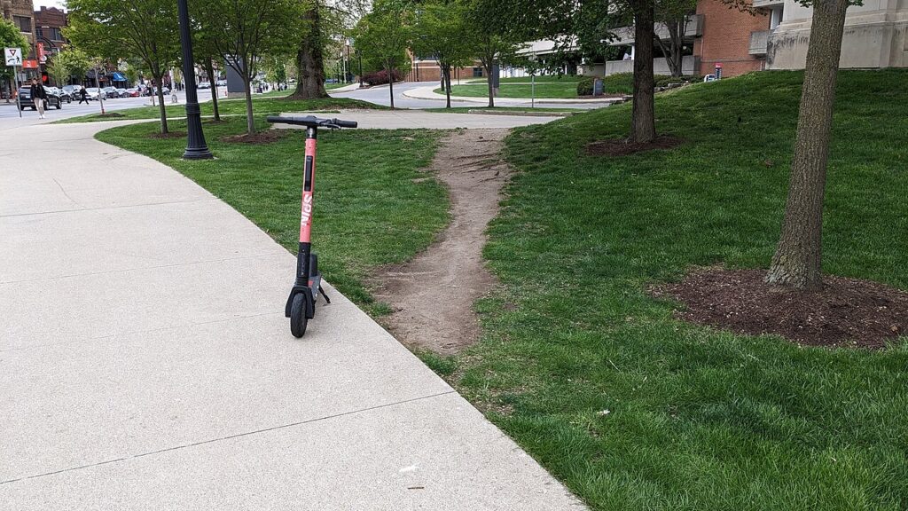 A dirt path, or "desire path," branches off from a sidewalk through grass and trees to another sidewalk, with a scooter positioned at the head of the desire path.