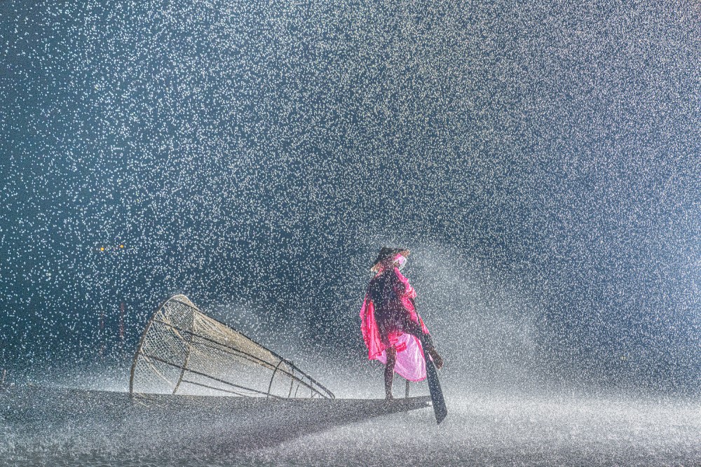 Photo "Resilience in the Rain" by Aung Chan Thar shows a person on a boat in a downpour, wearing a conical hat and a red raincoat and holding a paddle