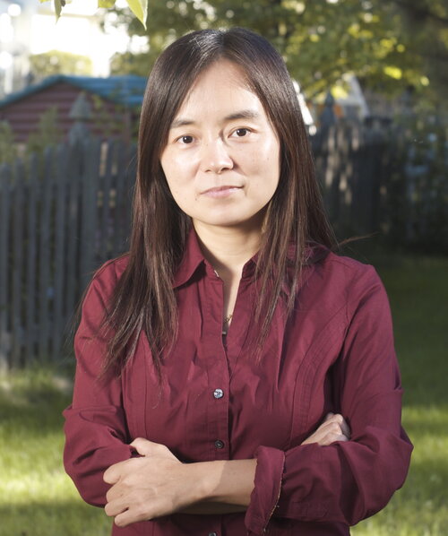 Zhuo Wang standing outside looking at the camera with her arms folded, with trees, a fence, and buildings in the background.