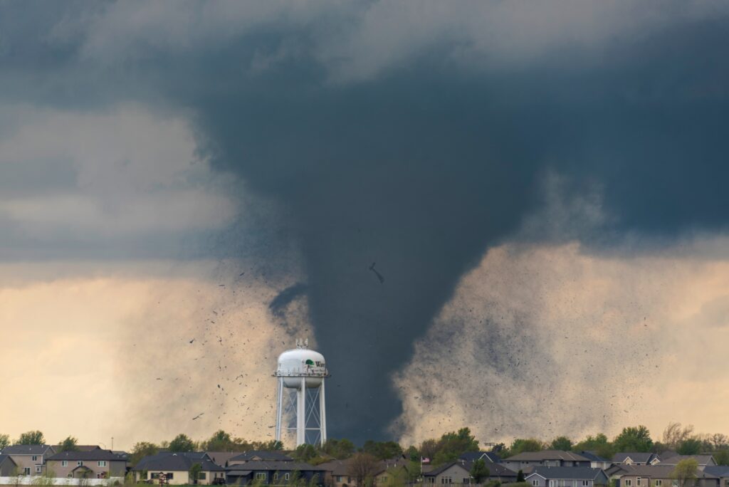 "Finger of God" by Caleb O'Gara. Photo of a tornado in the background, behind a town with a prominent water tower. The tornado is surrounded by a cloud of debris.