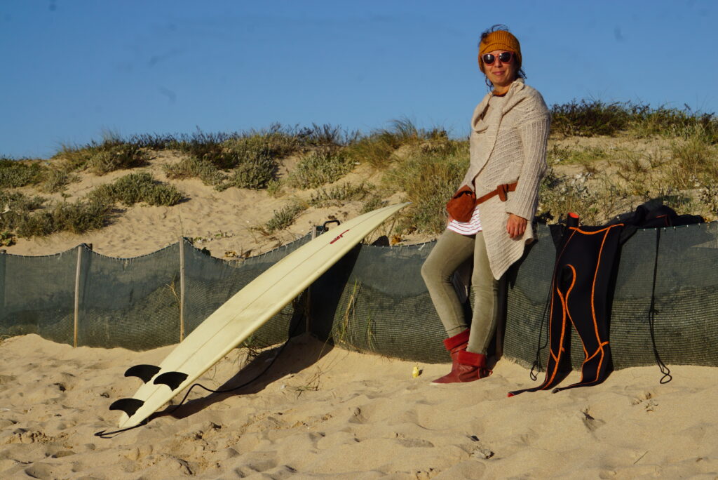 Desislava Petrova standing on a beach leaning against a low fence, with a surfboard next to her. She is facing the camera and smiling.