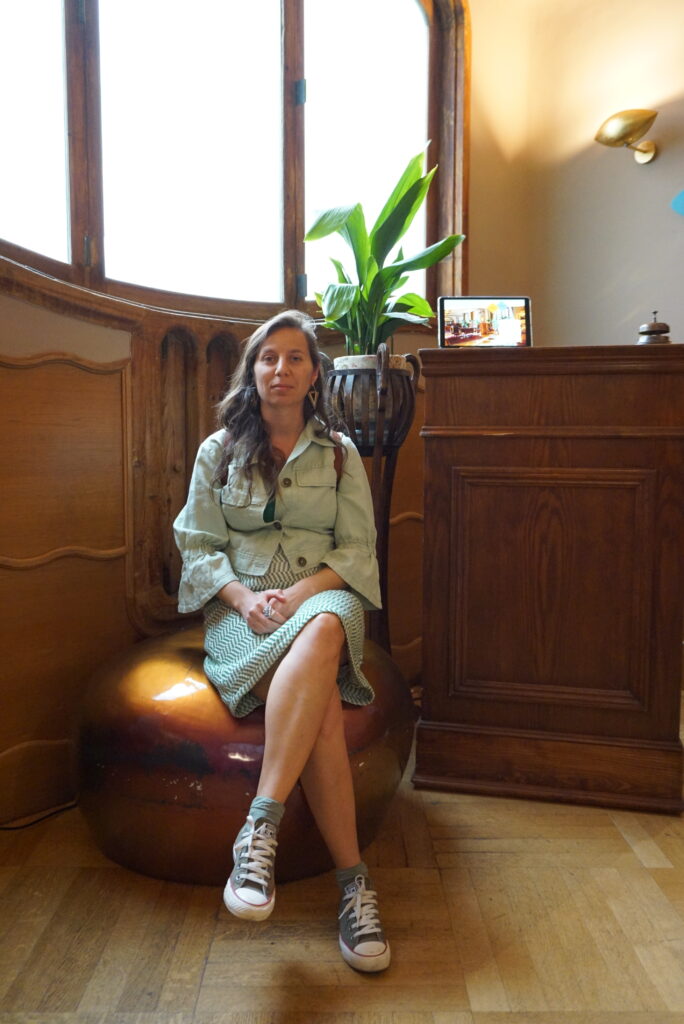 Desislava Petrova sitting and facing the camera in a room, with a plant directly behind her.