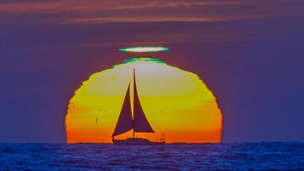 "Sailboat Sunset" by Jim Grant: Photo of a sailboat in front of the setting sun, with a green flash visible above the sun.