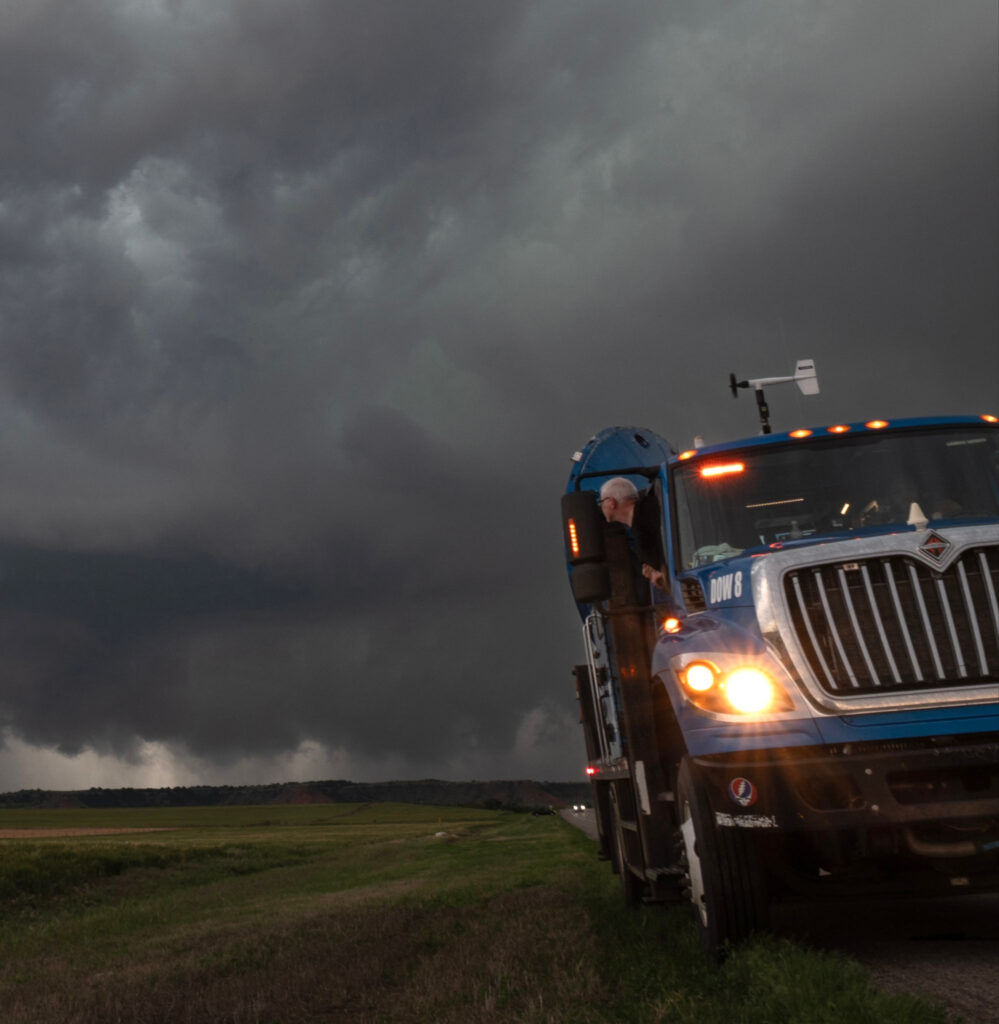 Photo: A large blue truck in the foreground with its lights on. A man leans out the window to look behind him at a large storm. Photo by Jen Walton, FARM Facility.