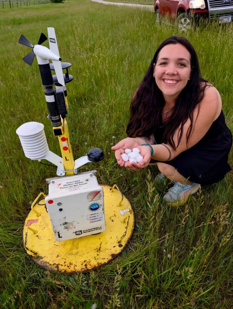 Photo of a smiling long-haired woman (Anastasia Tomanek) kneeling next to a yellow-painted metal Pod with multiple weather instruments attached. The woman is holding several large hailstones in her hands.