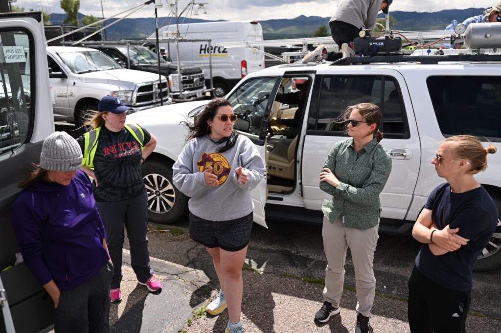 Photo of a group of people in casual clothes standing in front of white vehicles. A long-haired woman (Anastasia Tomanek) is talking to the others, who are watching her.