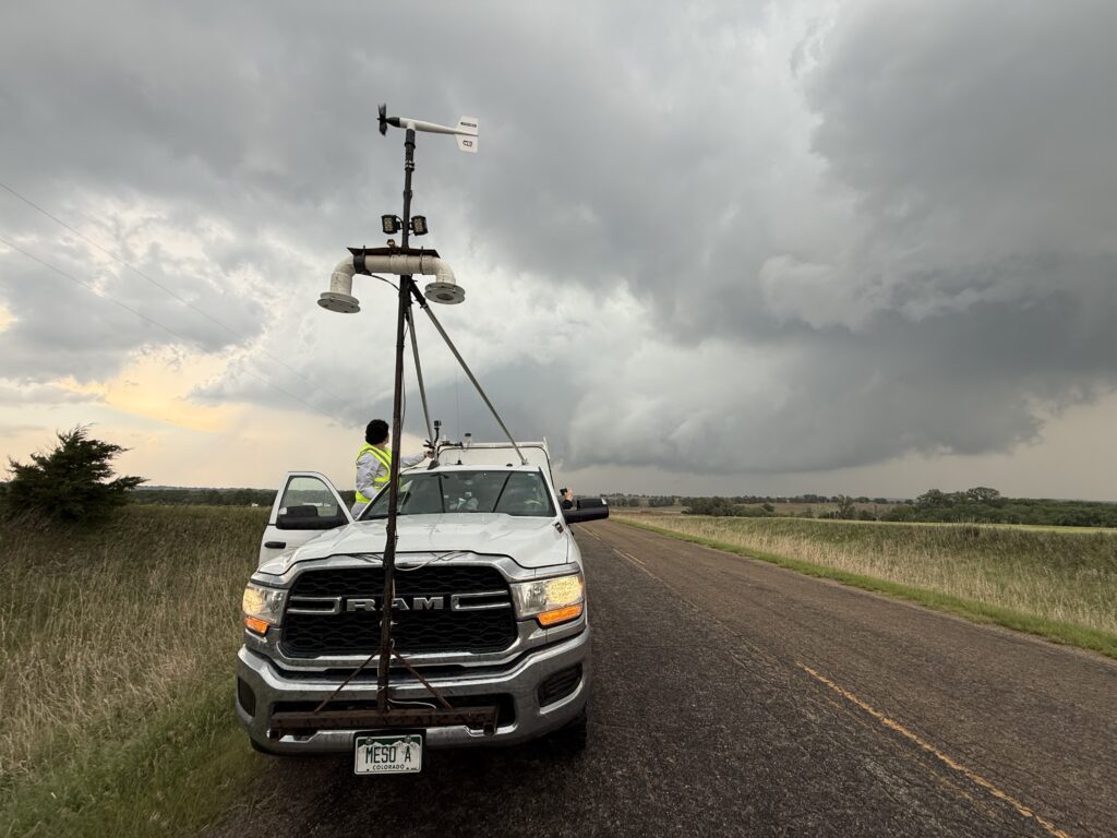 Photo of a white truck with meteorological instruments mounted above it. A woman is standing on the side of the truck, looking behind her at a storm.