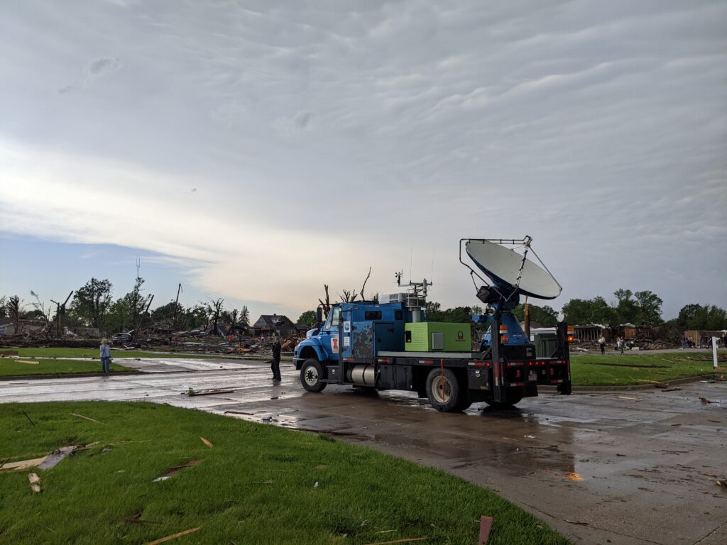 Photo: A blue flatbed truck with a radar mounted on its back, surrounded by debris, in front of several flattened buildings and downed trees.