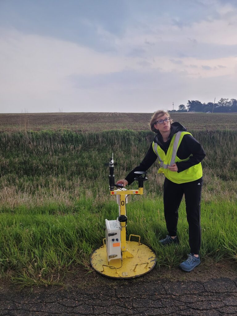 Photo of a woman (Jen Walton) in a safety vest next to a yellow metal Pod of meteorological instruments
