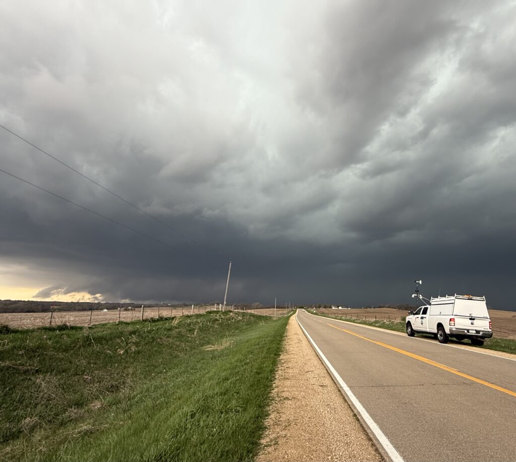 Photo of a white truck parked by the side of an empty road. The vehicle has meteorological instruments mounted on it. In the background, a large storm.