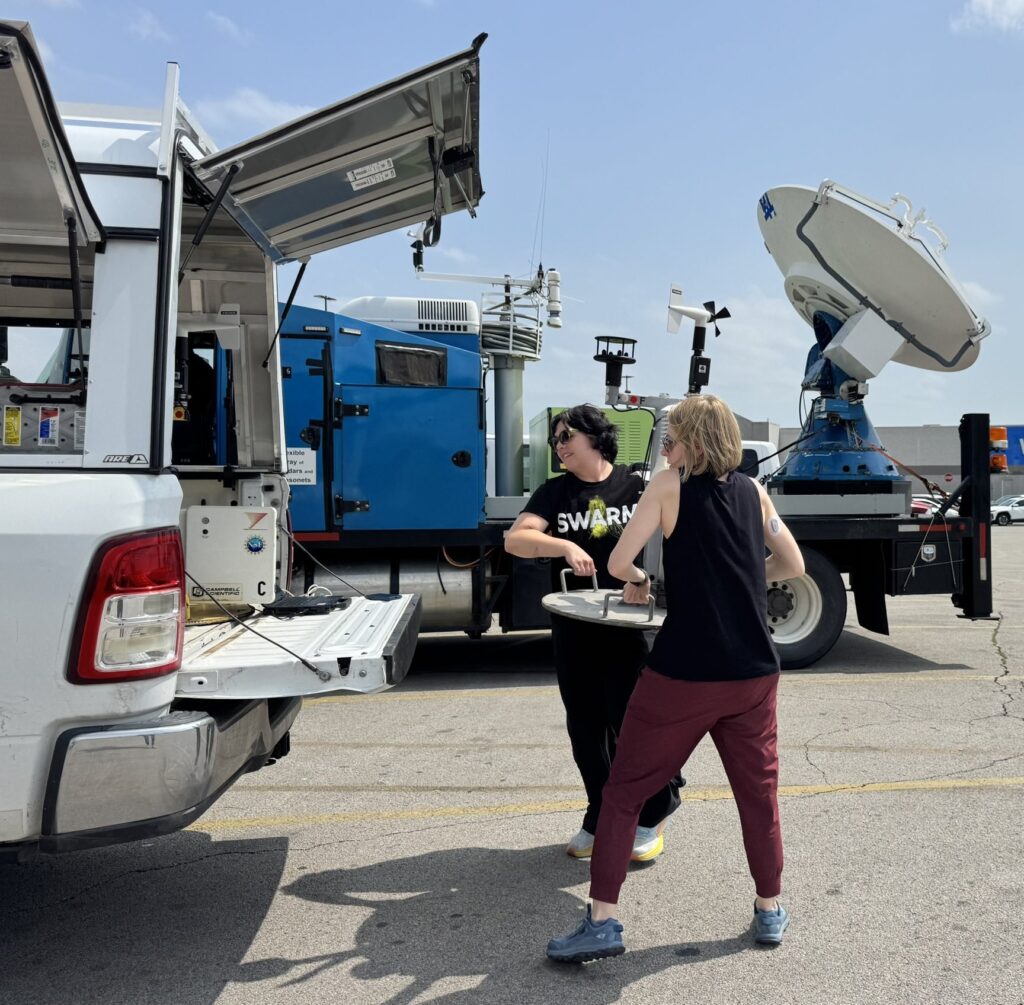 Photo of two women (Anastasia Tomanek and Jen Walton) working together to lift a Pod with meteorological instruments into the back of a truck.