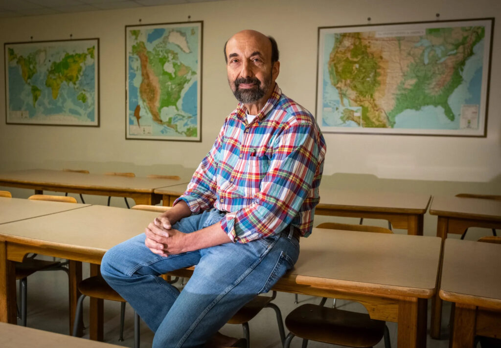 Walt Robinson sitting on a desk in a classroom, with maps on the wall in the background