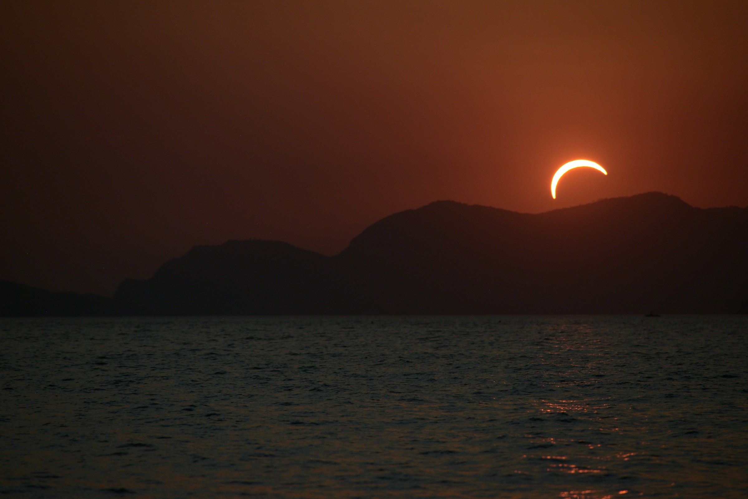 A partially eclipsed crescent sun above dark mountain silhouettes over a calm sea in Punta Bianca, Liguria, Italy on August 12, 2026.
