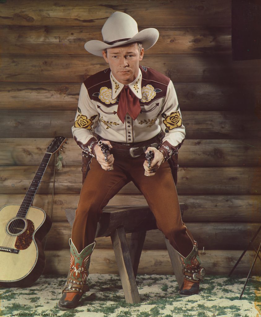 Roy Rogers standing in front of  wall holding a drawn pistol in each hand. A guitar is leaning against the wall. He is wearing a cowboy outfit, including a white hat.
